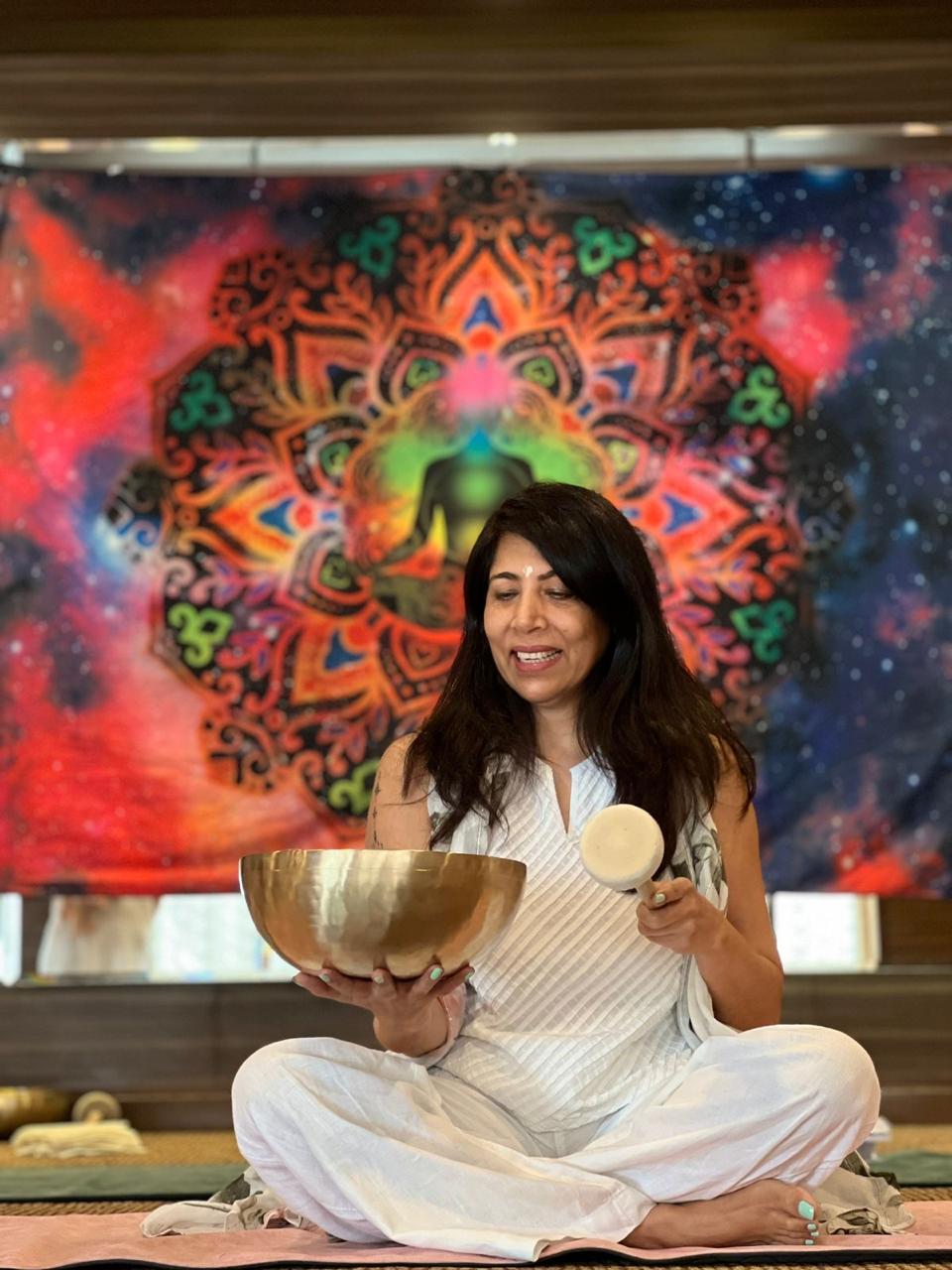 Shelly Rikhy Mandhar smiling warmly while holding a Tibetan singing bowl and mallet, seated in front of a vibrant mandala tapestry